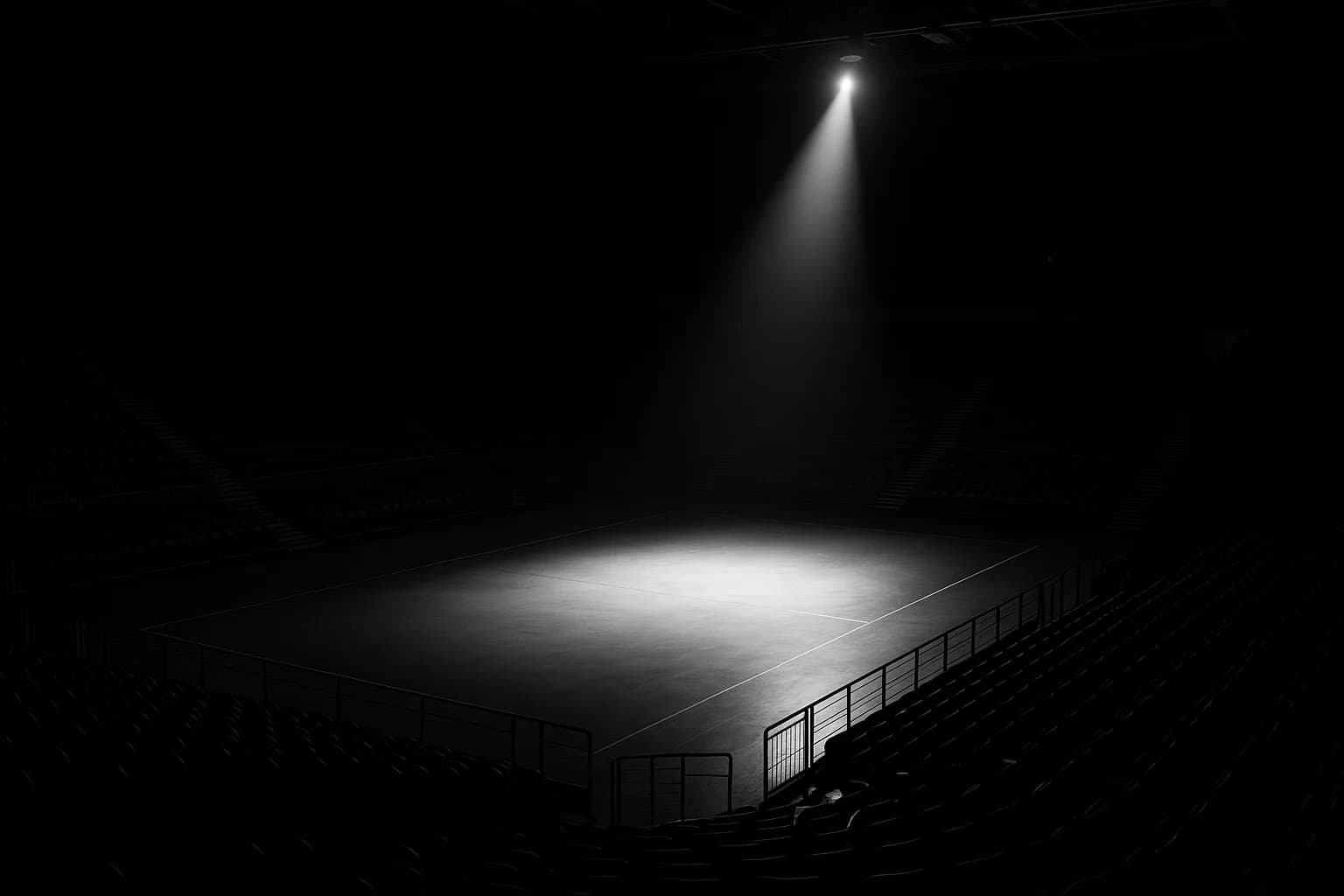 Schwarzweiß-Fotografie einer leeren modernen Indoor-Sportarena nach dem Event, einzelner Scheinwerfer beleuchtet die menschenleere Wettkampffläche von oben, ringsum dunkle Tribünenränge.